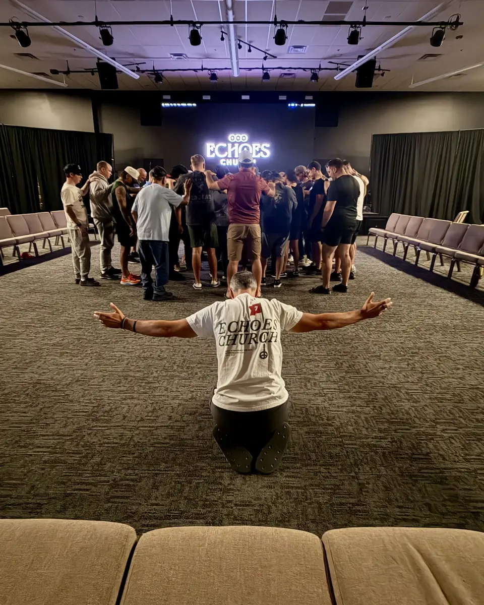 A man kneeling with arms open while a group gathers in a circle to pray